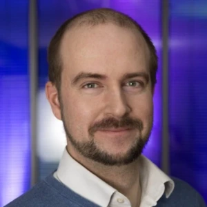 A smiling man with a short beard and mustache. He has short, dark hair. The man is wearing a blue sweater over a white shirt with an unbuttoned collar. Blurred violet and blue lights are visible in the background.