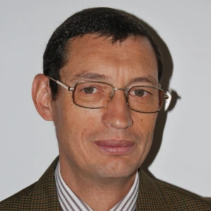 A man with short dark hair and glasses with thin metal frames. He is wearing a finely checkered brown and greenish blazer with a striped shirt. The man is looking at the camera with a subtle smile against a light background.