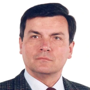 A man with short, neatly combed dark hair, looking directly at the camera. He is wearing a dark checkered blazer, a light shirt, and a dark red tie. The background of the photo is solid white.