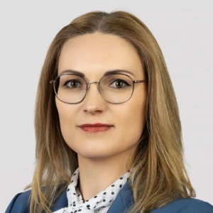 A woman with long, straight blonde hair and round, thin-framed glasses. She has a gentle smile and is wearing a blue blazer and a white blouse with black polka dots. The background is a neutral, light gray.