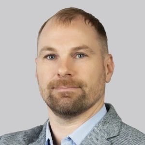 A man with short hair and a neatly trimmed brown beard and mustache. He is wearing a gray checkered blazer and a light blue shirt. The background of the photo is a neutral gray.