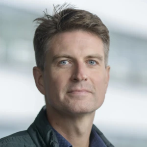 A light-eyed man with short, slightly messy brown hair. He is looking at the camera with a neutral facial expression, wearing a dark green jacket or blazer and a bluish shirt. The background is professionally blurred.
