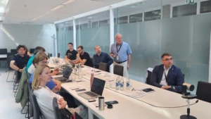 A professional meeting in a bright, modern glass-walled conference room. A man stands at the head of a long table presenting to a group of seated colleagues who are listening and working on laptops.