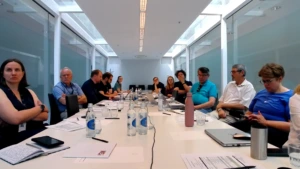 A group of professionals seated around a long white conference table during a meeting. The room features a modern design with glass walls, a white ceiling with a long recessed light panel, and water bottles and laptops spread across the table.
