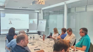 A presentation during a meeting in a conference room. A projector screen displays a slide titled "Research Programme Surfaces and Materials" with a presenter's photo, while participants sit along a long white table looking toward the screen.