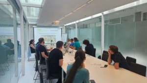 An angled view of a long white conference table filled with participants in a modern office building. A projector screen at the far end displays a presentation titled "Universidade de Aveiro - Facts and Figures."