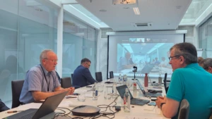 A close-up side perspective of attendees at a conference table. An older man in a blue button-down shirt talks at the left side of the table, while a large projector screen in the background shows a live video conference with multiple participants.