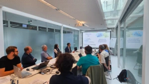 Attendees seated along a conference table watching a presentation displayed on a large screen at the front of the room. The slide title reads "Solutions for Self-Assembled Nanophotonics" by Nanoversa.