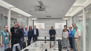 A group portrait of about fifteen professionals standing and posing inside a conference room in front of a projector screen showing a video call.