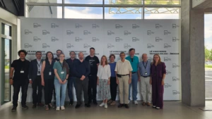 A group photo of twelve people standing in a line in front of a white promotional backdrop featuring repeating logos and the Lithuanian text "UŽ KIEKVIENOS TECHNOLOGIJOS &ndash; ŽMOGUS".