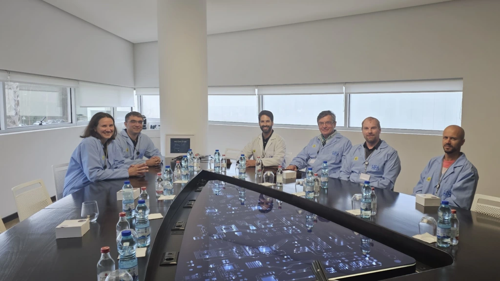 Six people wearing light blue lab coats sitting around a large conference table with a built-in illuminated circuit board display screen and bottles of water.