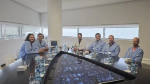 Six people wearing light blue lab coats sitting around a large conference table with a built-in illuminated circuit board display screen and bottles of water.