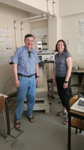 A man in a blue striped shirt and a woman in a grey polo shirt smiling while standing in front of laboratory scientific equipment.