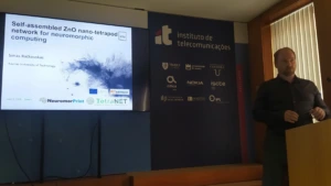 Simas secondment_1 Simas Račkauskas presenting a talk titled "Self-assembled ZnO nano-tetrapod network for neuromorphic computing" standing at a podium next to a presentation slide display.