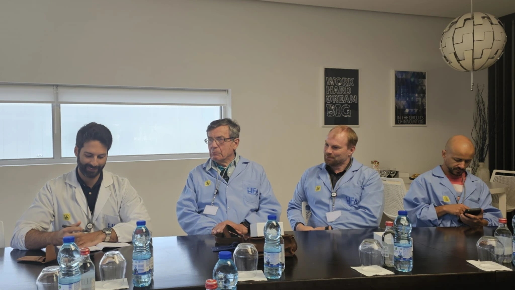 Four men wearing light blue lab coats seated side-by-side at a conference table with water bottles and glasses placed in front of them.