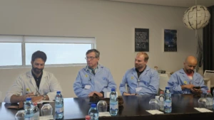 Simas secondment_2 Four men wearing light blue lab coats seated side-by-side at a conference table with water bottles and glasses placed in front of them.