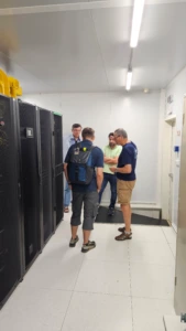 Tomas secondment_1 Four men standing in a modern server room, engaged in conversation next to tall racks of server cabinets.