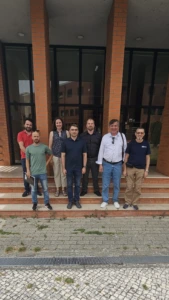 Tomas secondment_3 A group of seven researchers and professionals posing together in front of the brick entrance of an Instituto de Telecomunicações building.